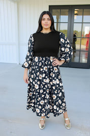 A woman wearing a black midi dress with a white and beige floral print, featuring a solid black ribbed bodice and voluminous three-quarter sleeves, paired with silver heels.