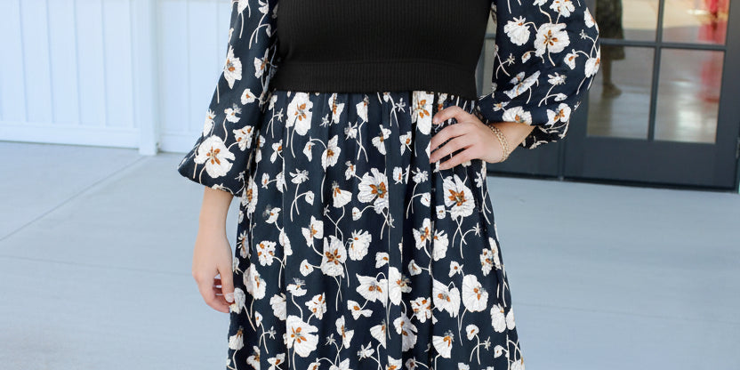A woman wearing a black midi dress with a white and beige floral print, featuring a solid black ribbed bodice and voluminous three-quarter sleeves, paired with silver heels.
